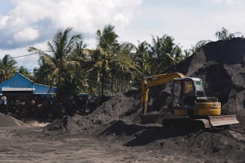A yellow excavator is positioned on a large pile of dark soil in a yard surrounded by tall palm trees. Next to it, a blue structure with a sloped roof is visible, alongside a stack of weathered barrels. The sky is partly cloudy, and the scene conveys an industrial setting amidst a tropical environment.