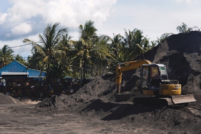 A yellow excavator is positioned on a large pile of dark soil in a yard surrounded by tall palm trees. Next to it, a blue structure with a sloped roof is visible, alongside a stack of weathered barrels. The sky is partly cloudy, and the scene conveys an industrial setting amidst a tropical environment.
