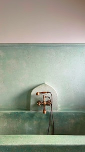A rustic faucet fixture mounted against a textured, speckled green wall over a matching green stone or concrete basin. The design has a vintage or classical feel, with brass elements and a simple aesthetic.