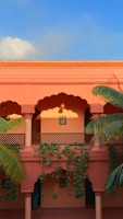 A two-story building with a warm terracotta facade, featuring traditional arches and decorative lattice work. Tropical plants with green leaves climb along the walls and columns. The upper level showcases framed artwork, and there is a clear blue sky above, imparting a serene, inviting atmosphere.