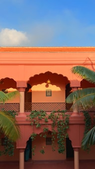 A two-story building with a warm terracotta facade, featuring traditional arches and decorative lattice work. Tropical plants with green leaves climb along the walls and columns. The upper level showcases framed artwork, and there is a clear blue sky above, imparting a serene, inviting atmosphere.