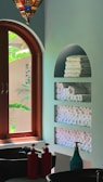 An interior space featuring a wooden arched window with lush green foliage visible outside. Inside, there is a wall with built-in shelves holding neatly stacked and rolled white towels. A decorative hanging light fixture with colorful glass pieces is visible above. Two sinks are present with red and turquoise soap dispensers on the counter.