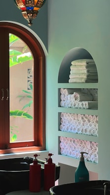 An interior space featuring a wooden arched window with lush green foliage visible outside. Inside, there is a wall with built-in shelves holding neatly stacked and rolled white towels. A decorative hanging light fixture with colorful glass pieces is visible above. Two sinks are present with red and turquoise soap dispensers on the counter.