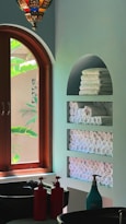 An interior space featuring a wooden arched window with lush green foliage visible outside. Inside, there is a wall with built-in shelves holding neatly stacked and rolled white towels. A decorative hanging light fixture with colorful glass pieces is visible above. Two sinks are present with red and turquoise soap dispensers on the counter.