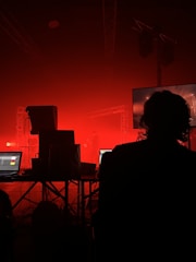 A dark room with a prominent red lighting gives an intense atmosphere. Silhouetted figures are seated in front of computer equipment and monitors, possibly controlling audio or visual aspects of a performance. Stage lights and structural trusses are visible, indicating a live event setup.