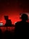 A dark room with a prominent red lighting gives an intense atmosphere. Silhouetted figures are seated in front of computer equipment and monitors, possibly controlling audio or visual aspects of a performance. Stage lights and structural trusses are visible, indicating a live event setup.