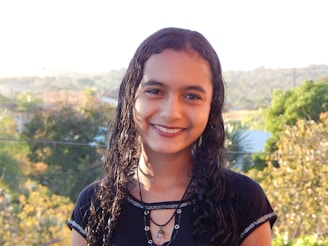 A person with wet, curly hair smiles against a natural background of blurred trees and foliage. The lighting is bright, suggesting it's a sunny day.