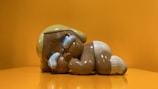 A glossy, stylized sculpture of a character is resting on its side against a vibrant orange background. The character has exaggerated features with a large head, oversized limbs, and a serene expression. Its colors include earthy browns and muted blues, and it appears to be made of a shiny material.