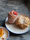 A delicious assortment of pastries including a tall, spiral pastry dusted with sugar and topped with a red center, a rounded pastry covered in almond slices, and a small pastry with a slightly burnt edge. They are presented on two white plates with blue rims placed on a dark, textured stone surface.
