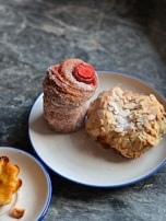 A delicious assortment of pastries including a tall, spiral pastry dusted with sugar and topped with a red center, a rounded pastry covered in almond slices, and a small pastry with a slightly burnt edge. They are presented on two white plates with blue rims placed on a dark, textured stone surface.