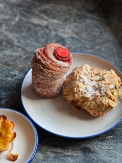 A delicious assortment of pastries including a tall, spiral pastry dusted with sugar and topped with a red center, a rounded pastry covered in almond slices, and a small pastry with a slightly burnt edge. They are presented on two white plates with blue rims placed on a dark, textured stone surface.