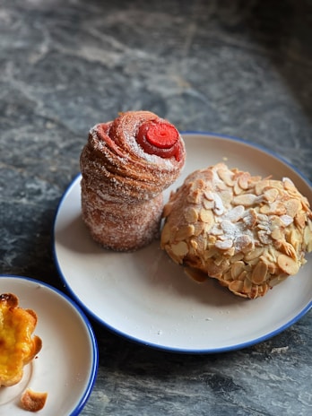 A delicious assortment of pastries including a tall, spiral pastry dusted with sugar and topped with a red center, a rounded pastry covered in almond slices, and a small pastry with a slightly burnt edge. They are presented on two white plates with blue rims placed on a dark, textured stone surface.