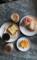 A marble table with an assortment of pastries and beverages. One plate has layered pastries with a red topping and sliced almonds. Another plate contains a loaf of sweet bread with a wooden spoon. A small plate holds a tart with a glossy top. Two jars contain iced coffee and a chocolate drink.