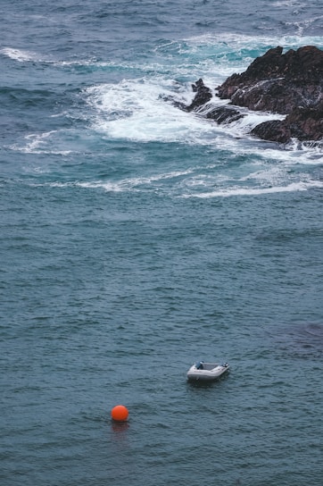 A small inflatable boat is moored in the ocean near a rocky shoreline. The water is a deep blue with white waves breaking over the rocks. An orange buoy floats nearby, adding a splash of color to the scene.