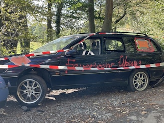 A parked black station wagon with a damaged appearance, surrounded by red and white warning tape. Graffiti in red paint is visible on the vehicle's side. The car is situated in a wooded area, with trees and greenery in the background.