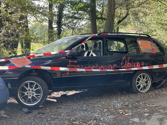 A parked black station wagon with a damaged appearance, surrounded by red and white warning tape. Graffiti in red paint is visible on the vehicle's side. The car is situated in a wooded area, with trees and greenery in the background.