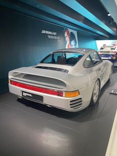 A classic white Porsche sports car is showcased in a museum setting. The car is positioned on a shiny black floor with a backdrop featuring motivational text in both German and English. A vintage poster of individuals is visible on the wall, adding a nostalgic atmosphere.