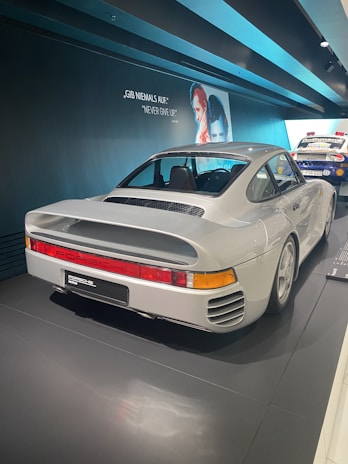A classic white Porsche sports car is showcased in a museum setting. The car is positioned on a shiny black floor with a backdrop featuring motivational text in both German and English. A vintage poster of individuals is visible on the wall, adding a nostalgic atmosphere.