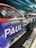 A close-up view of a race car featuring prominent branding and sponsorship logos. The car has a sleek, aerodynamic design with a colorful livery in blue, red, and green hues. The text includes 'PAUL', 'DEKRA', and 'DTM', indicating sponsorship or team details. The car is exhibited in a modern showroom or display area, as suggested by the lighting and setting.