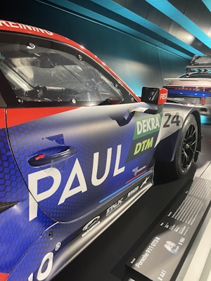 A close-up view of a race car featuring prominent branding and sponsorship logos. The car has a sleek, aerodynamic design with a colorful livery in blue, red, and green hues. The text includes 'PAUL', 'DEKRA', and 'DTM', indicating sponsorship or team details. The car is exhibited in a modern showroom or display area, as suggested by the lighting and setting.