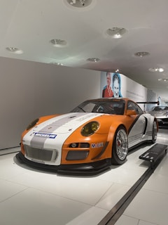 A sleek, orange and white race car is displayed in a modern indoor exhibit. The car, featuring prominent Michelin and Mobil1 logos, is positioned at an angle showcasing its front and side view. The background reveals a sterile, clean environment with white walls and ceiling fixtures. A large photograph of a smiling person is also visible on the wall behind the car.