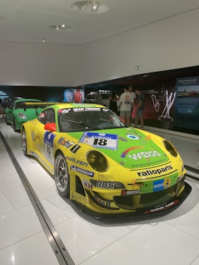 A brightly colored race car with a yellow and green livery is displayed indoors. It features various sponsor logos, including BBS, Falken, and Mobil 1. The car is positioned on a shiny white floor with track lighting overhead. In the background, another car and several people viewing the display can be observed.