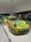 A brightly colored race car with a yellow and green livery is displayed indoors. It features various sponsor logos, including BBS, Falken, and Mobil 1. The car is positioned on a shiny white floor with track lighting overhead. In the background, another car and several people viewing the display can be observed.