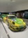 A brightly colored race car with a yellow and green livery is displayed indoors. It features various sponsor logos, including BBS, Falken, and Mobil 1. The car is positioned on a shiny white floor with track lighting overhead. In the background, another car and several people viewing the display can be observed.