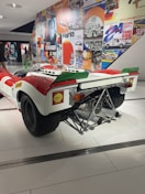 A vintage racing car displayed in a museum setting, featuring a white and red color scheme with a prominent exhaust system and branding stickers like Shell. The background includes a wall covered with vibrant, retro-themed posters showcasing racing events and automotive brands.