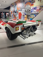 A vintage racing car displayed in a museum setting, featuring a white and red color scheme with a prominent exhaust system and branding stickers like Shell. The background includes a wall covered with vibrant, retro-themed posters showcasing racing events and automotive brands.