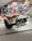 A vintage racing car displayed in a museum setting, featuring a white and red color scheme with a prominent exhaust system and branding stickers like Shell. The background includes a wall covered with vibrant, retro-themed posters showcasing racing events and automotive brands.