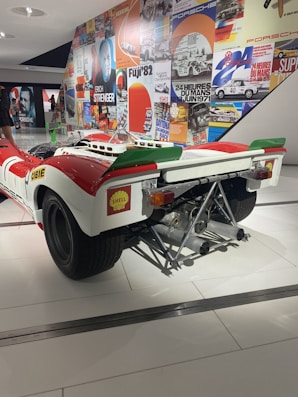 A vintage racing car displayed in a museum setting, featuring a white and red color scheme with a prominent exhaust system and branding stickers like Shell. The background includes a wall covered with vibrant, retro-themed posters showcasing racing events and automotive brands.