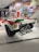 A vintage racing car displayed in a museum setting, featuring a white and red color scheme with a prominent exhaust system and branding stickers like Shell. The background includes a wall covered with vibrant, retro-themed posters showcasing racing events and automotive brands.