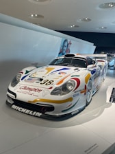 A race car is displayed in a well-lit, modern exhibition space. The car has colorful decals, including sponsor logos such as Michelin and BOSCH, and the number 38 on its hood. The car has a streamlined design, a front splitter, and a large rear wing. The background includes a white wall and some overhead light fixtures, with part of another race car visible in the distance.