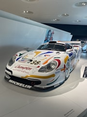 A race car is displayed in a well-lit, modern exhibition space. The car has colorful decals, including sponsor logos such as Michelin and BOSCH, and the number 38 on its hood. The car has a streamlined design, a front splitter, and a large rear wing. The background includes a white wall and some overhead light fixtures, with part of another race car visible in the distance.