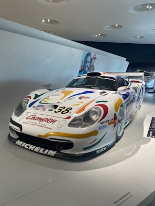A race car is displayed in a well-lit, modern exhibition space. The car has colorful decals, including sponsor logos such as Michelin and BOSCH, and the number 38 on its hood. The car has a streamlined design, a front splitter, and a large rear wing. The background includes a white wall and some overhead light fixtures, with part of another race car visible in the distance.