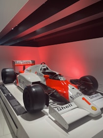 A vintage Formula 1 racing car with prominent red and white Marlboro and Shell branding is on display under a red light. The car features large black tires, a sleek aerodynamic body, and the number 1 displayed prominently on the front. The exhibit is set in a modern, minimalistic environment, enhancing the car's vibrant colors.