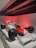 A vintage Formula 1 racing car with prominent red and white Marlboro and Shell branding is on display under a red light. The car features large black tires, a sleek aerodynamic body, and the number 1 displayed prominently on the front. The exhibit is set in a modern, minimalistic environment, enhancing the car's vibrant colors.