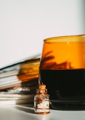 A small glass vial filled with white crystals is adorned with copper wire and positioned in front of a large amber glass cup filled with a dark liquid. Soft lighting casts warm reflections and shadows, enhancing the amber color.