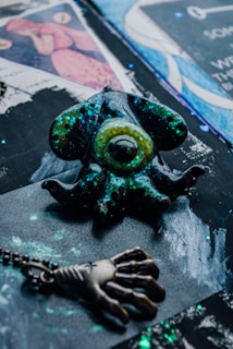 A decorative object with a design resembling an eye surrounded by tentacles, featuring a shiny, speckled surface with hues of green and blue. A pendant with a hand-like design is also present, connected by a chain. The background consists of abstract patterns and colors, including blues, black, and purples.