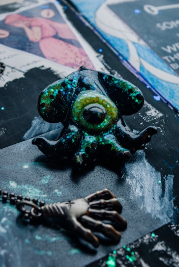 A decorative object with a design resembling an eye surrounded by tentacles, featuring a shiny, speckled surface with hues of green and blue. A pendant with a hand-like design is also present, connected by a chain. The background consists of abstract patterns and colors, including blues, black, and purples.