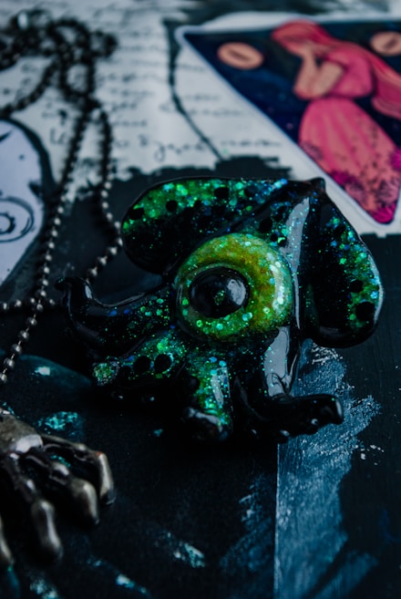 A close-up of a metallic octopus pendant with a chain, covered in green and black glitter. There's a tarot card in the background featuring a figure in a red dress, and text or drawings on a textured surface.