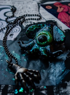 An abstract scene featuring a dark, glossy pendant in the shape of an squid-like creature, adorned with iridescent green and blue glitter and attached to a metal chain that ends in a claw. The background consists of black paper, ornate designs, and a drawing of a figure in a pink dress.