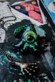 A mystical pendant shaped like an eye surrounded by octopus tentacles, coated with glittering shades of green and black. Nearby, a metal chain with a skeletal hand charm adds a gothic touch. The background features artistic elements including handwritten script and an image of a woman in red inside a triangle.