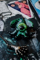 A mystical pendant shaped like an eye surrounded by octopus tentacles, coated with glittering shades of green and black. Nearby, a metal chain with a skeletal hand charm adds a gothic touch. The background features artistic elements including handwritten script and an image of a woman in red inside a triangle.