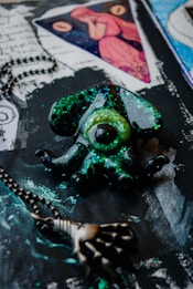 A mystical pendant shaped like an eye surrounded by octopus tentacles, coated with glittering shades of green and black. Nearby, a metal chain with a skeletal hand charm adds a gothic touch. The background features artistic elements including handwritten script and an image of a woman in red inside a triangle.