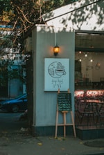A cozy cafe corner is depicted with a small sign featuring a coffee cup and slice of cake illustration. Below, a menu board lists various coffee options. Soft, warm lighting from a hanging lantern enhances the relaxed ambiance. The exterior has trees casting shadows, creating a blend of natural and urban elements.