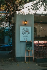 A cozy cafe corner is depicted with a small sign featuring a coffee cup and slice of cake illustration. Below, a menu board lists various coffee options. Soft, warm lighting from a hanging lantern enhances the relaxed ambiance. The exterior has trees casting shadows, creating a blend of natural and urban elements.