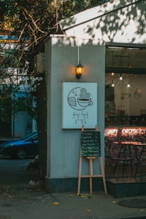 A cozy cafe corner is depicted with a small sign featuring a coffee cup and slice of cake illustration. Below, a menu board lists various coffee options. Soft, warm lighting from a hanging lantern enhances the relaxed ambiance. The exterior has trees casting shadows, creating a blend of natural and urban elements.