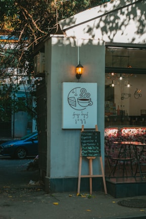 A cozy cafe corner is depicted with a small sign featuring a coffee cup and slice of cake illustration. Below, a menu board lists various coffee options. Soft, warm lighting from a hanging lantern enhances the relaxed ambiance. The exterior has trees casting shadows, creating a blend of natural and urban elements.